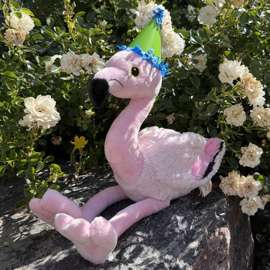 A pink plush flamingo with a green party hat sits on a rock amid white flowers and foliage, ready for DZCA animal care support as part of the 130th Anniversary Adoption Bundle.