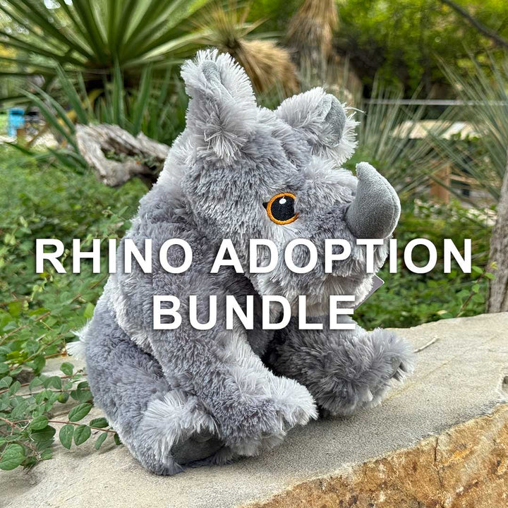 A plush rhinoceros toy rests on a stone ledge outdoors among green plants. Bold white text reads: Rhino Adoption Bundle—help protect wildlife with every Rhino Adoption Bundle purchase.