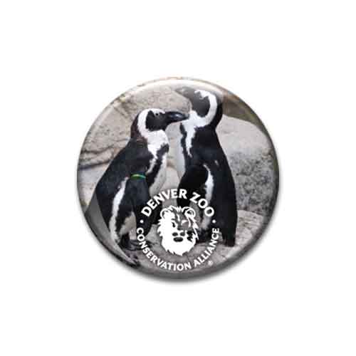 Two penguins face each other in front of rocks, with a DZCA Penguin Glass Dome Magnet featuring the Conservation Alliance logo and a lion illustration overlaid at the bottom of the image.