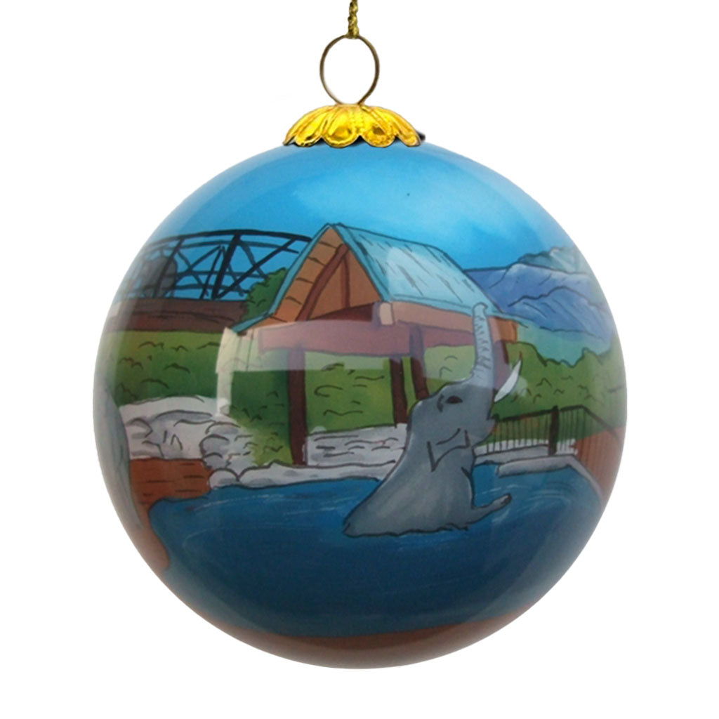 The Denver Zoo Elephant Ornament features a round design with an elephant in a pool, surrounded by a zoo scene with a bridge, greenery, and building beneath a shiny blue sky.