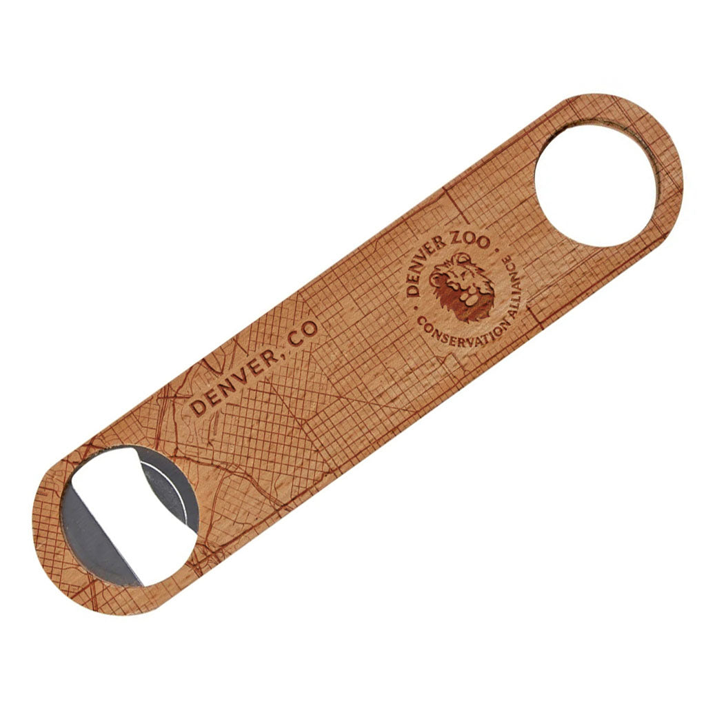 The DZCA Bottle Opener is a flat wooden barware piece engraved with a map, “DENVER, CO,” and the Denver Zoo Conservation lion logo. It features a metal insert on one end and a circular hole on the other.