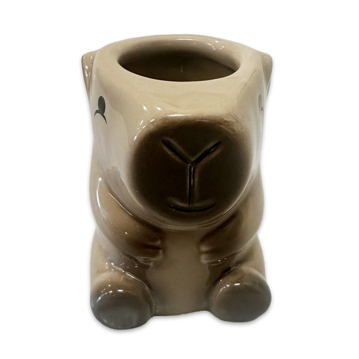The Capybara 3D Shot Glass is a small, light brown ceramic vessel shaped like a sitting capybara with black eyes, round ears, and a subtle smile. Its open top lets you use it as a quirky novelty shot glass. Background is white.