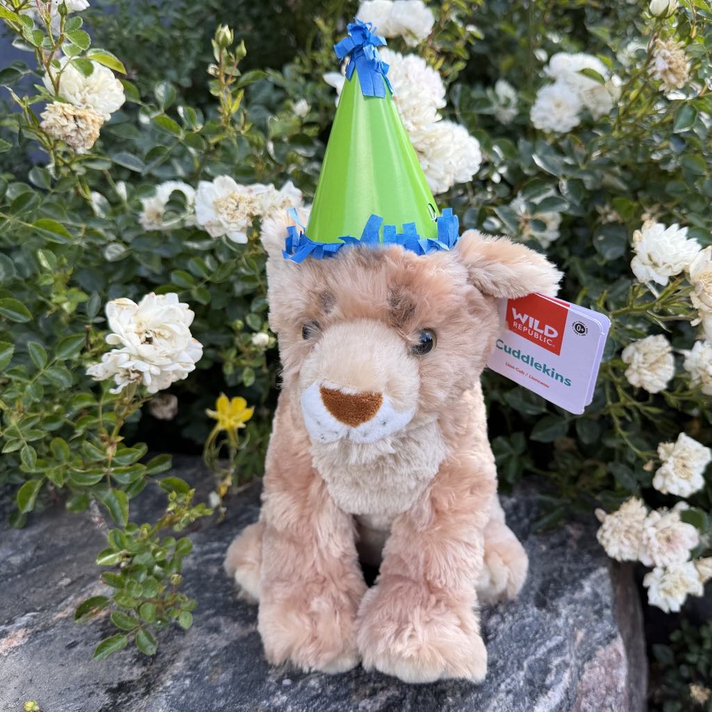 A plush lion toy from the 130th Anniversary Adoption Bundle, wearing a green party hat with blue trim, sits on a rock amid blooming white flowers, supporting animal care through the DZCA plush adoption program.