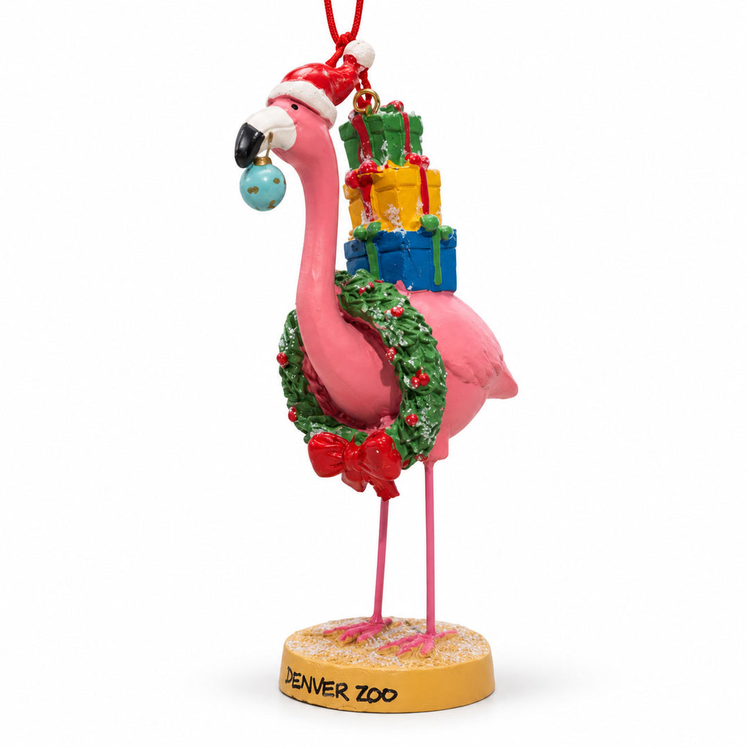A DZCA Flamingo Wreath Ornament wearing a Santa hat and holiday wreath, carrying gift boxes on its back while holding a small ornament in its beak, standing on a base labeled Denver Zoo.