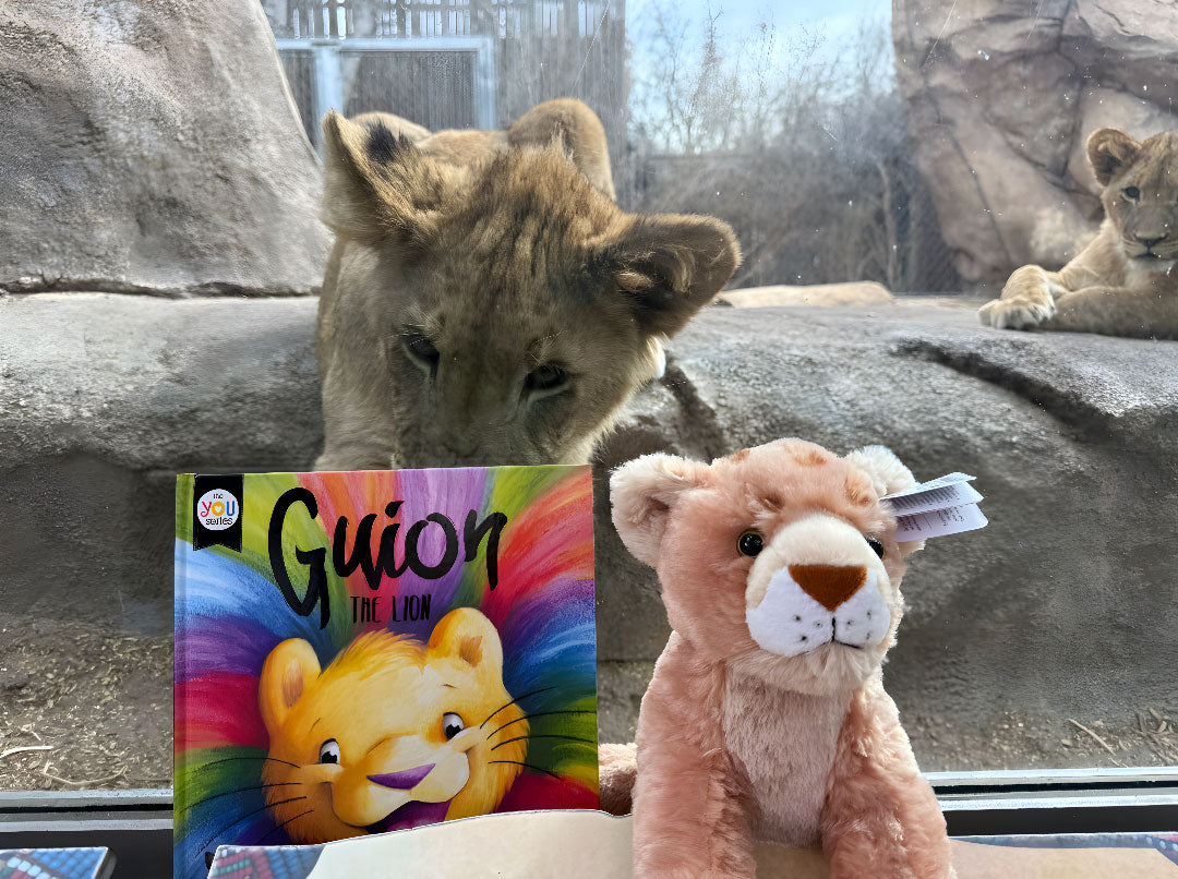 An African lion cub looks through glass behind the Guion the Lion Adoption Bundle—a set featuring the Guion the Lion children’s book and a light brown lion plush—displayed at a zoo enclosure. Adopt this bundle today!.