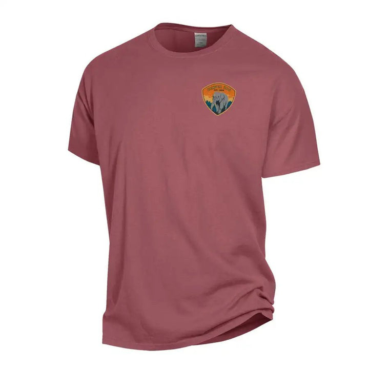 The Denver Zoo Elephant Patch T-Shirt is a mauve short-sleeve, ringspun cotton shirt featuring a small, colorful patch with an elephant and scenery on the left chest; the rest of the shirt is plain with no other visible designs.