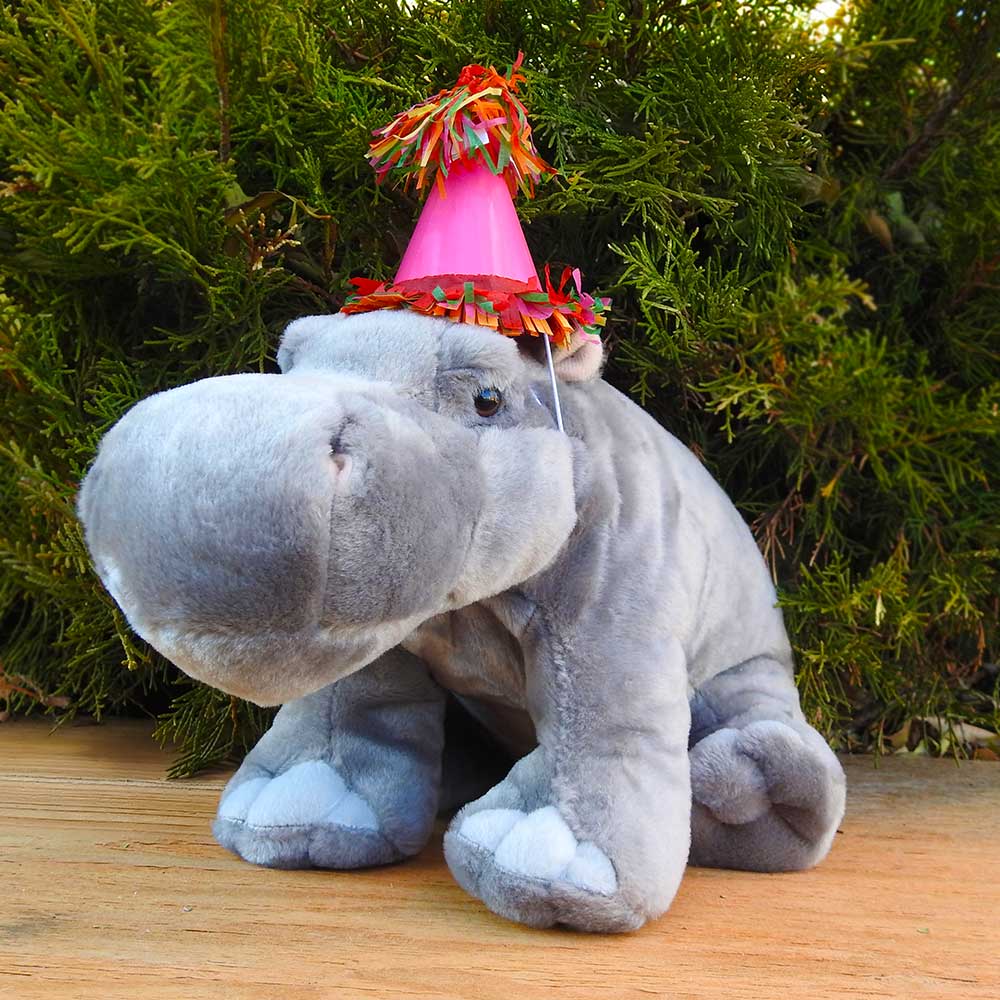 A gray plush hippo in a pink party hat with colorful fringe sits on a wooden surface, surrounded by green foliage—a charming part of our Birthday Adoption Bundle, perfect for adopting or gifting a special plush animal.