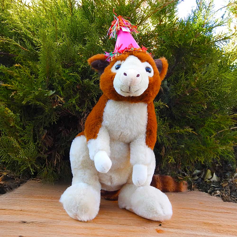 A brown and white plush horse from the Birthday Adoption Bundle sits on a wooden surface outdoors, sporting a colorful party hat. Perfect for an Adopt an Animal celebration, green bushes create a lively background.