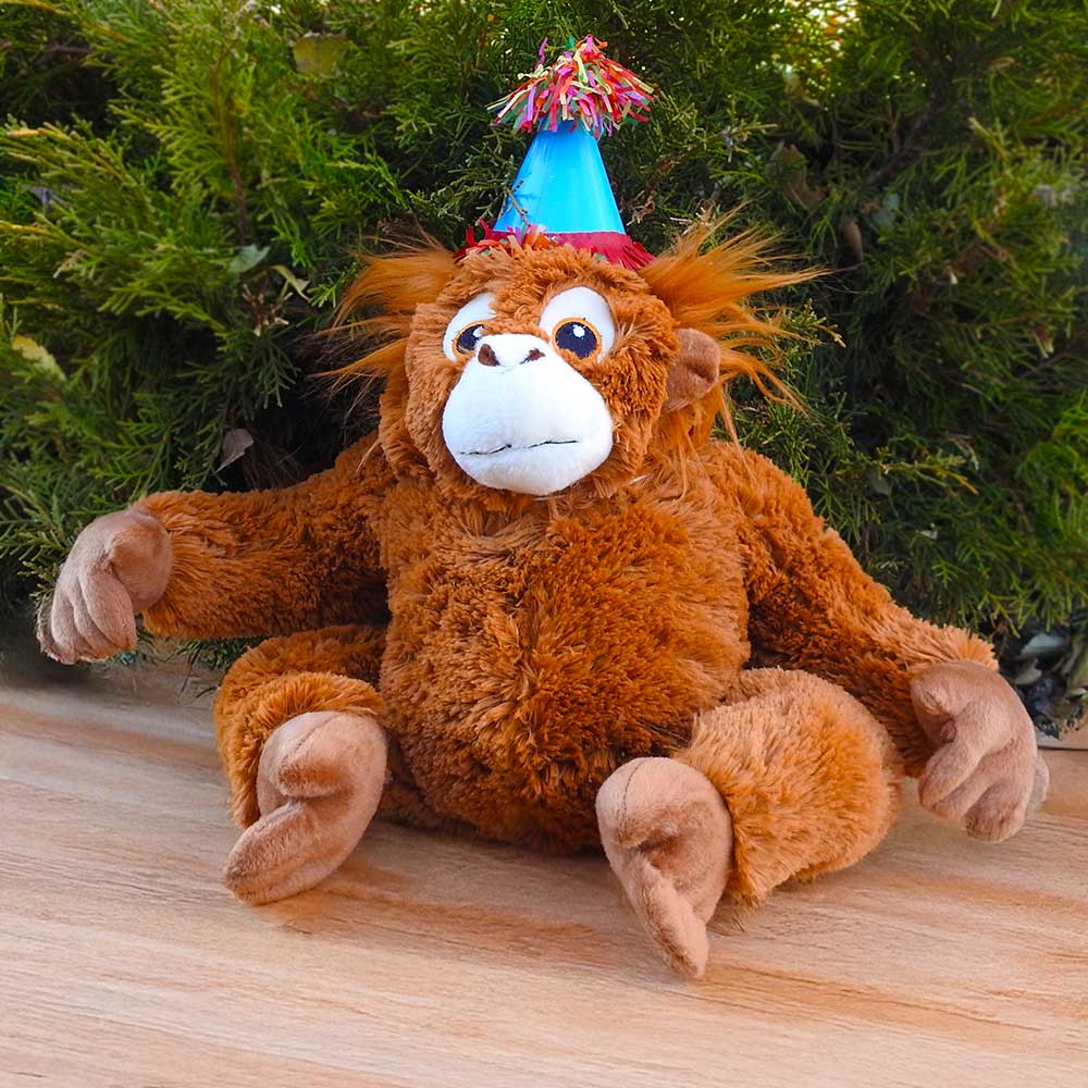 A plush monkey with a blue party hat and colorful tassels sits on wood before green bushes, ready to join your Birthday Adoption Bundle celebration.