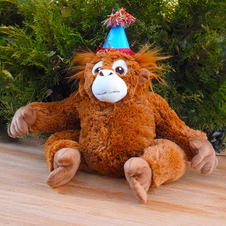A plush monkey with a blue party hat and colorful tassels sits on wood before green bushes, ready to join your Birthday Adoption Bundle celebration.