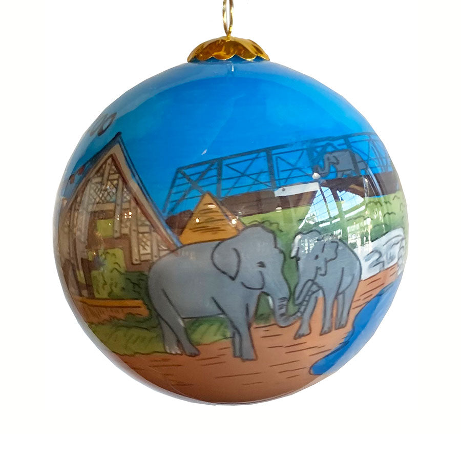 The Denver Zoo Elephant Ornament is a round blue ornament showing elephants painted on sandy ground by water, with buildings and a bridge in the background.