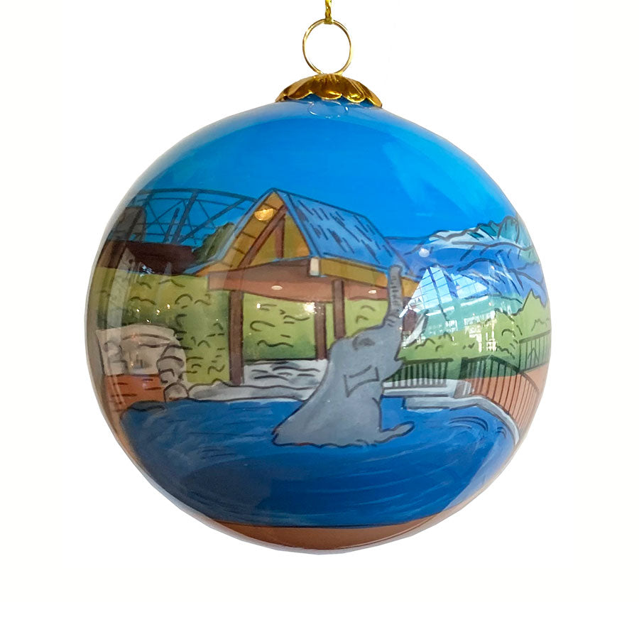 A painted Denver Zoo Elephant Ornament shows an elephant at the zoo with a wooden shelter, trees, and a fence in the background under a bright blue sky.