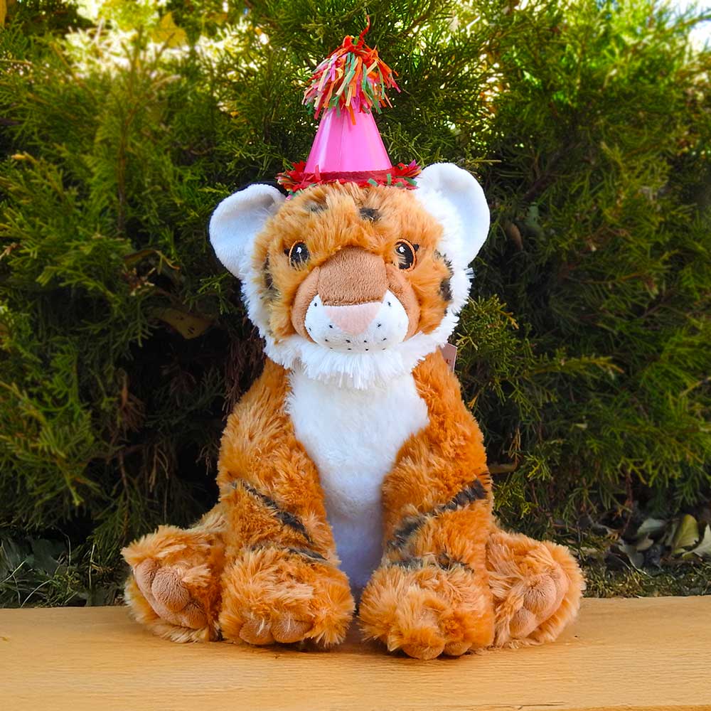 A plush tiger toy wearing a pink party hat with tassels sits on a wooden surface outdoors, surrounded by green bushes—an adorable part of the Birthday Adoption Bundle, perfect for celebrating special moments.