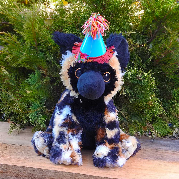 A plush African wild dog with a blue party hat and colorful fringe sits on wood by green bushes—perfect for the Birthday Adoption Bundle.